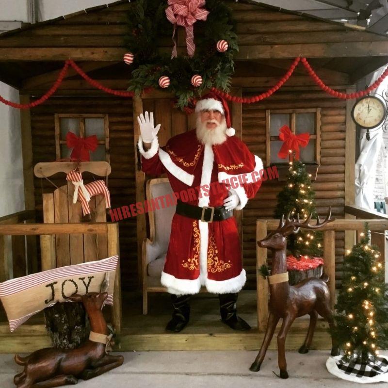 Real bearded Santa for hire in Port St. Lucie waving from a log cabin porch beside a carved reindeer.