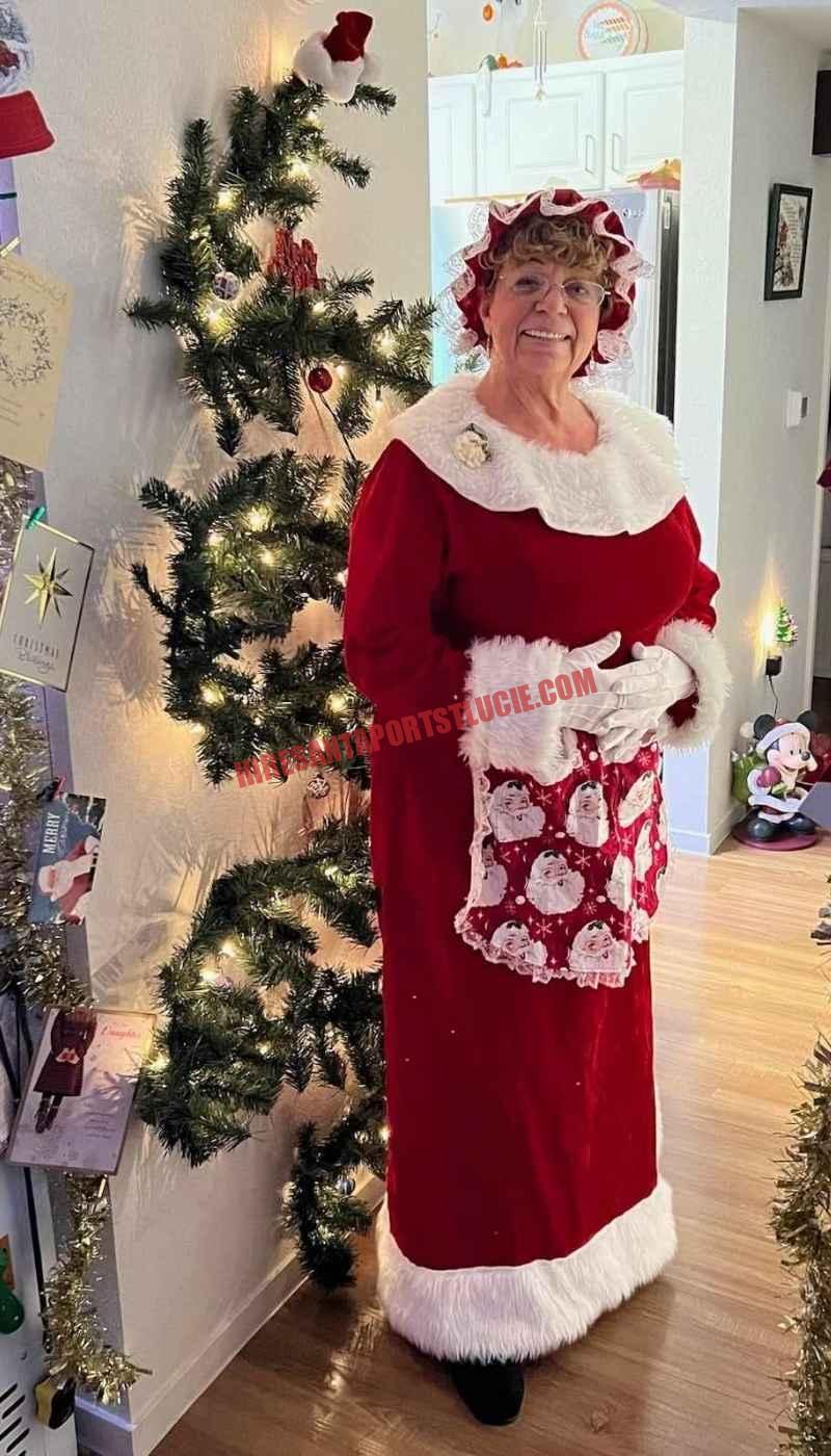 Mrs. Claus for hire in Port St. Lucie standing in a hallway beside a slim lit tree in a red dress and apron.