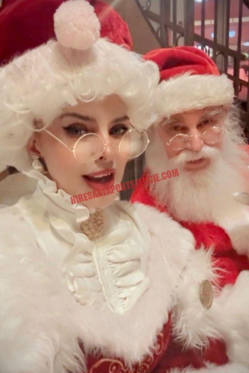 About Hire Santa Port St. Lucie: Mrs. Claus in white ruffles standing beside Santa in a red suit.